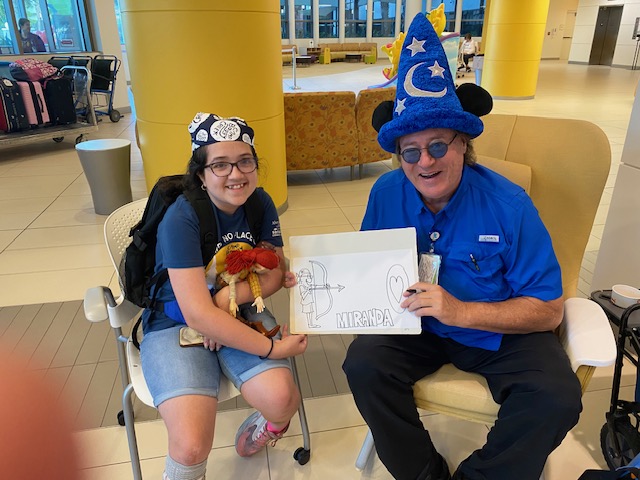 Girl sitting on chair holding puppet and man with sorcerer hat sitting on chair both jointly holding a certificate.