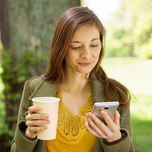 Young woman relaxing in a park with a coffee and a mobile phone reading a newsletter